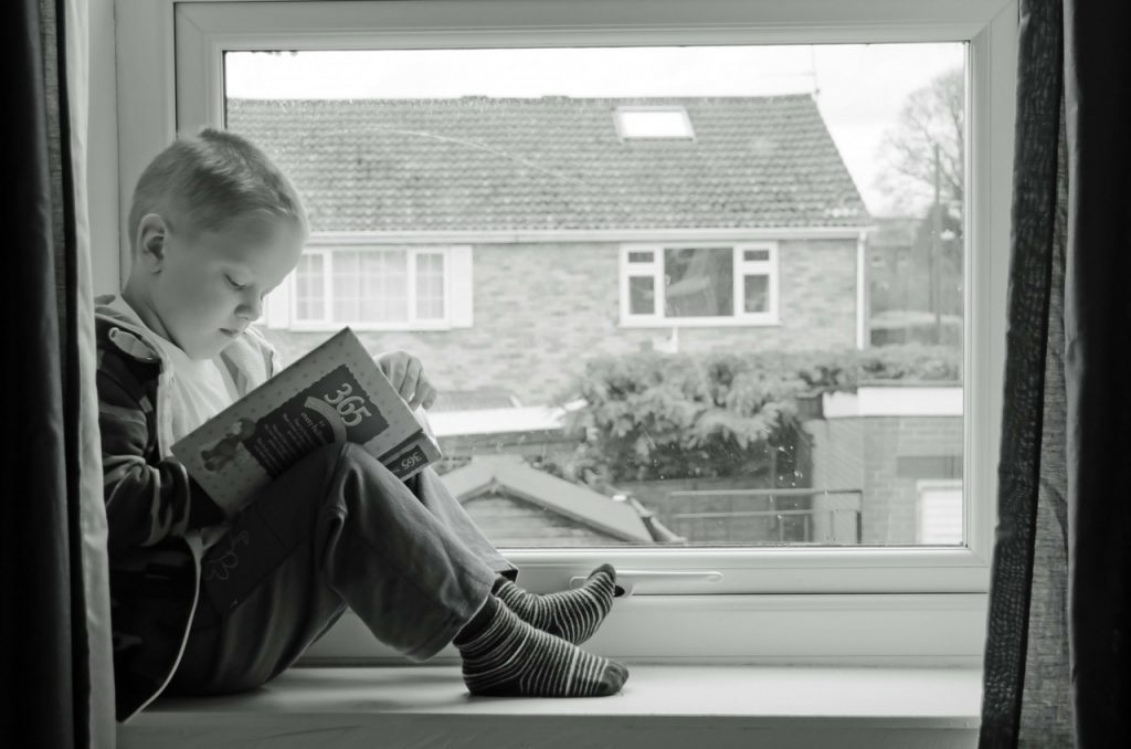 A kid reading story book sitting on a window sill 